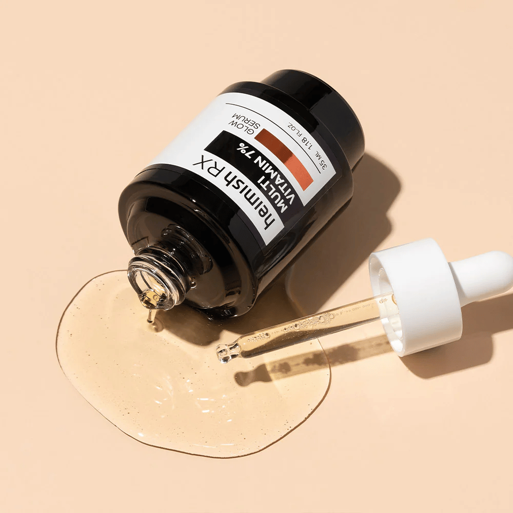 Heimish RX Multi Vitamin 7% Glow Serum adagiato su uno sfondo beige con siero sparso intorno alla bottiglia e una pipetta riempita di prodotto, che mette in risalto la sua formula ricca di nutrienti.