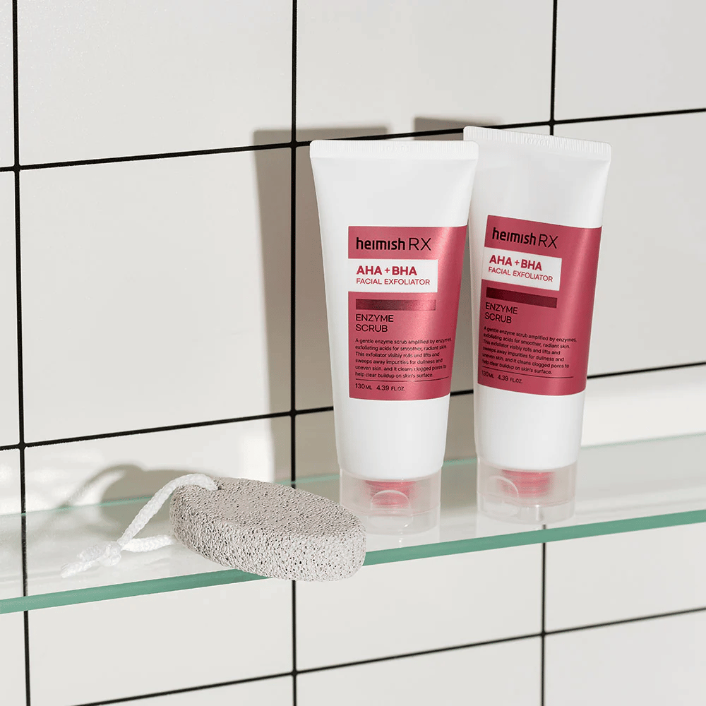 Due tubi Heimish RX AHA + BHA Esfoliante Viso posizionati su una mensola di vetro in un bagno con parete di piastrelle bianche. Accanto ai prodotti c'è una pietra pomice con un cordino bianco. Un'immagine di cura della pelle elegante e minimalista.