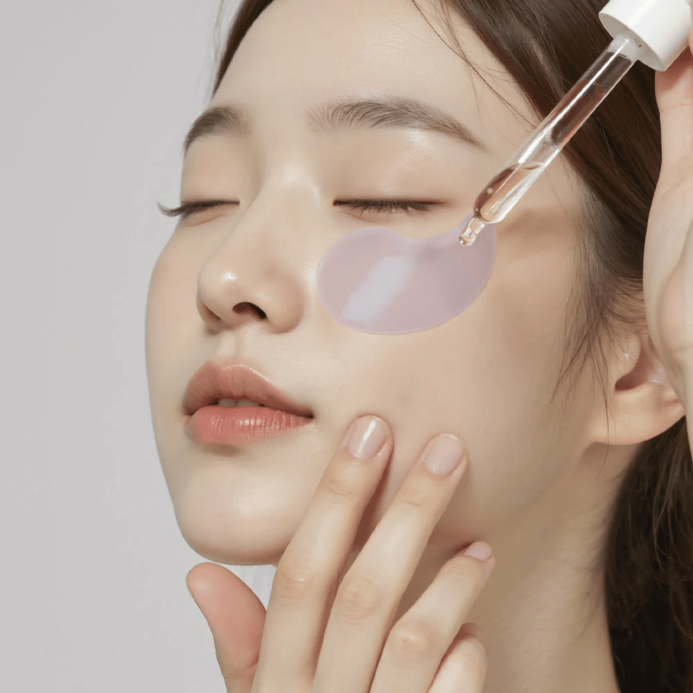 Una donna con un patch oculare in idrogel viola applica il siero sotto l'occhio con una pipetta. Il prodotto idrata e cura la pelle sensibile intorno agli occhi.