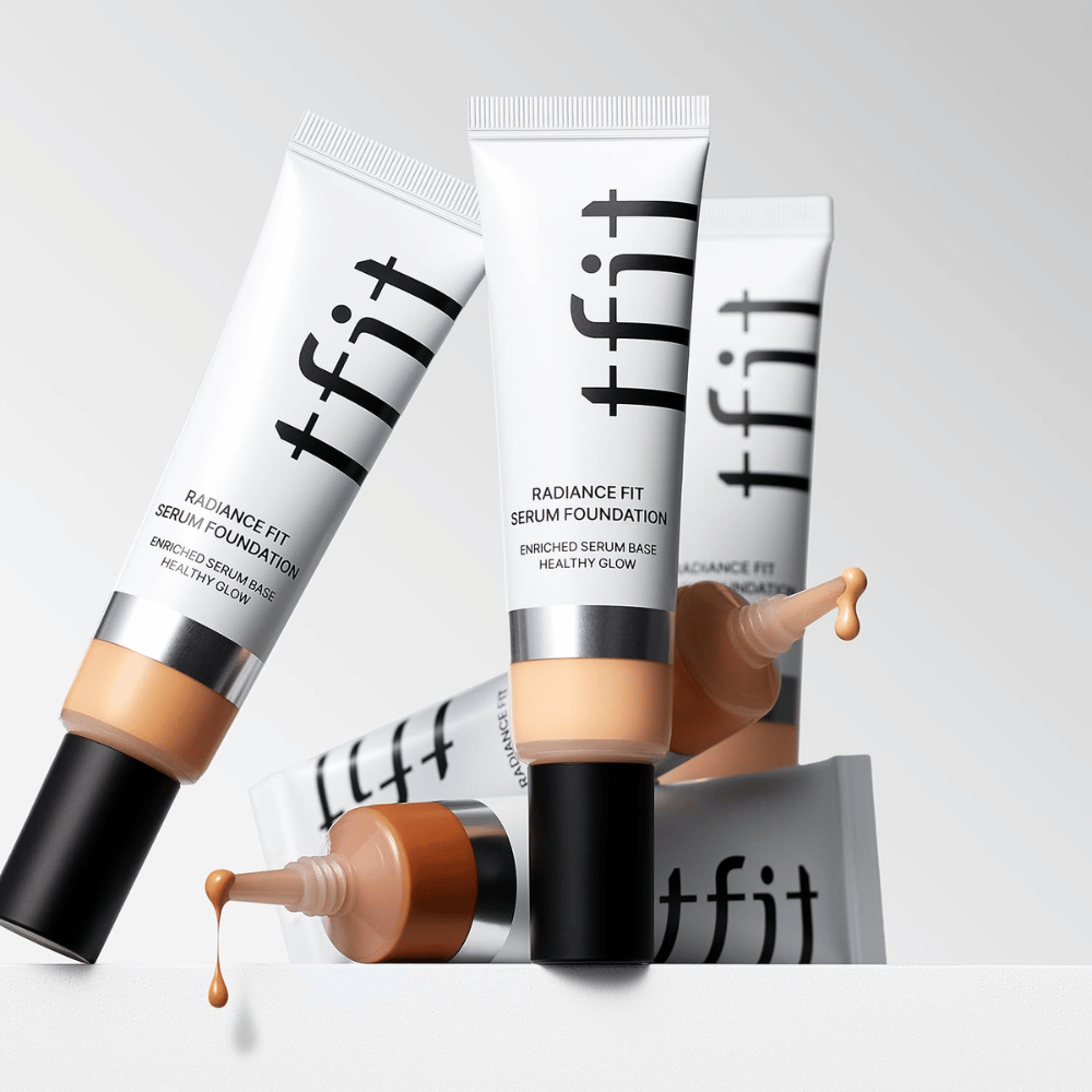 Diverse tubetti di TFIT Radiance Fit Serum Foundation in diverse tonalità, uno dei quali con fondotinta che gocciola, su sfondo grigio chiaro.