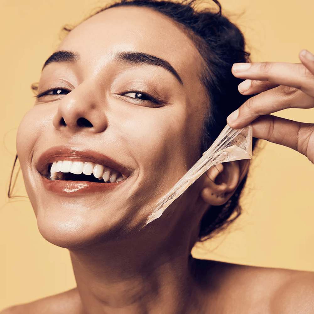 L'immagine mostra una donna con un grande sorriso che sta rimuovendo una maschera peel-off dal viso. La donna appare felice e soddisfatta mentre utilizza il prodotto di bellezza, il che trasmette una sensazione di benessere e cura della pelle.