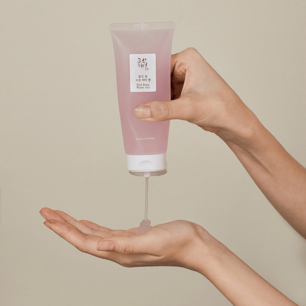L'immagine mostra una persona che tiene un tubo di "Red Bean Water Gel" da Beauty of Joseon e spremendo un gel trasparente nella sua mano. Il prodotto sembra essere destinato alla cura della pelle e ha una confezione rosa morbida e attraente.