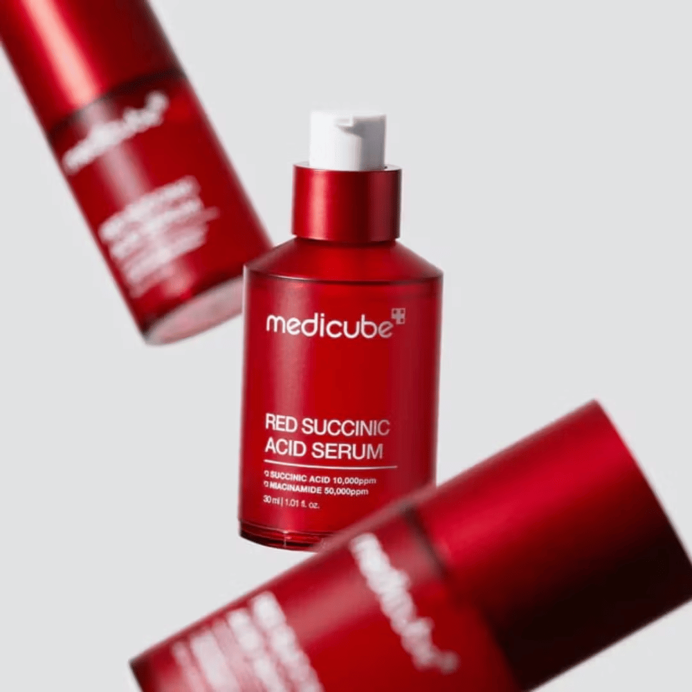 Medicube Red Succinic Acid Serum in bottiglia rossa con pompa, circondata da più bottiglie identiche su sfondo chiaro. Il prodotto contiene acido succinico e niacinamide per trattare acne e macchie pigmentarie.