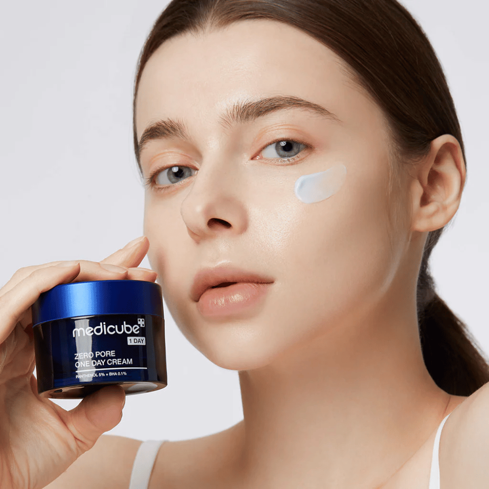 Donna con un look naturale tiene Medicube Zero Pore One Day Cream. Una piccola quantità di crema è applicata sulla sua guancia. Il prodotto è progettato per migliorare la struttura della pelle e minimizzare la dimensione dei pori, con una formula leggera e idratante.