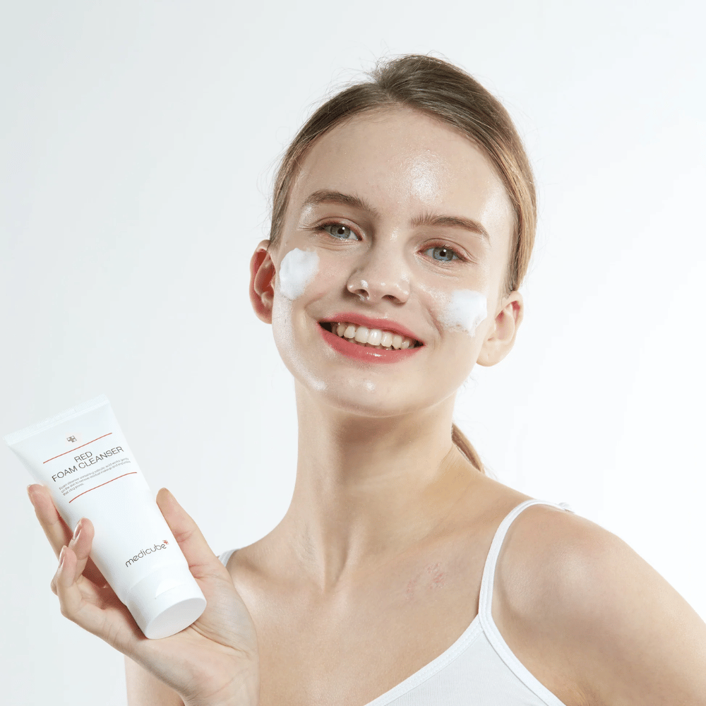 Giovane donna con schiuma detergente sul viso tiene un tubo di Medicube Red Foam Cleanser e sorride alla fotocamera. Indossa una canottiera bianca e lo sfondo è chiaro e semplice.