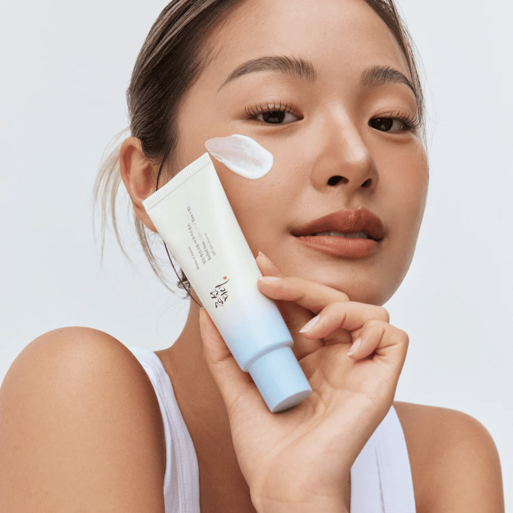 Donna tiene Beauty of Joseon Relief Sun: crema solare Aqua-Fresh Rice + B5 e mostra la sua consistenza cremosa sulla guancia. Un prodotto solare leggero che idrata e protegge la pelle con SPF 50+ PA++++.