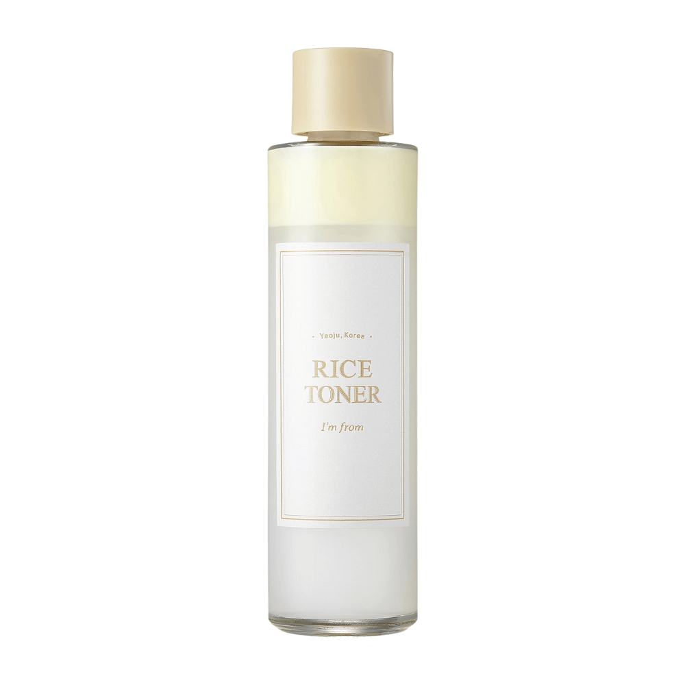 Una bottiglia trasparente con tappo dorato ed etichetta che indica "Rice Toner I'm from Yeojü, Corea". La bottiglia contiene un liquido chiaro, probabilmente tonico viso a base di estratto di riso. Il design è pulito e minimalista. Lo sfondo è bianco.