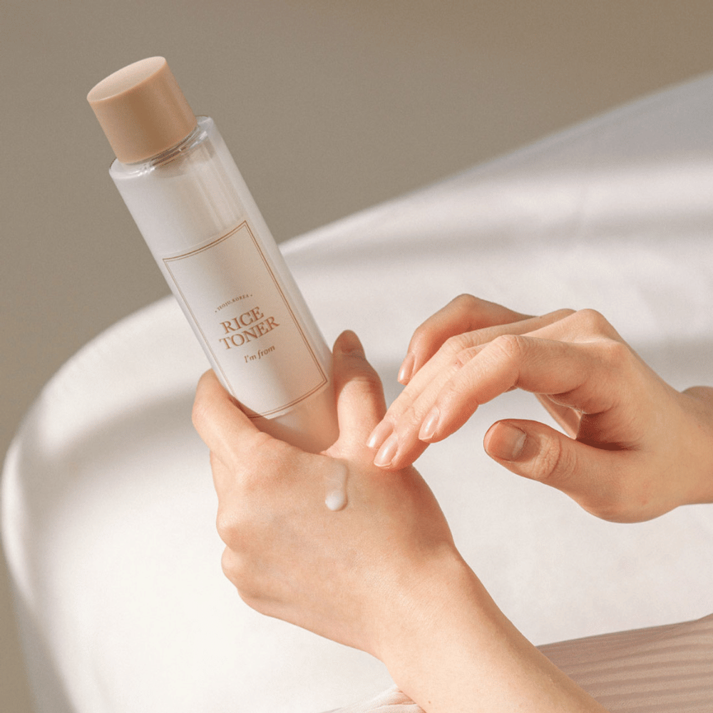 Mani che tengono una bottiglia di riso tonico viso etichettata "I'm from". Una goccia del prodotto è applicata su una mano mentre l'altra mano si prepara ad applicarla. La scena è illuminata dolcemente con un tono caldo.