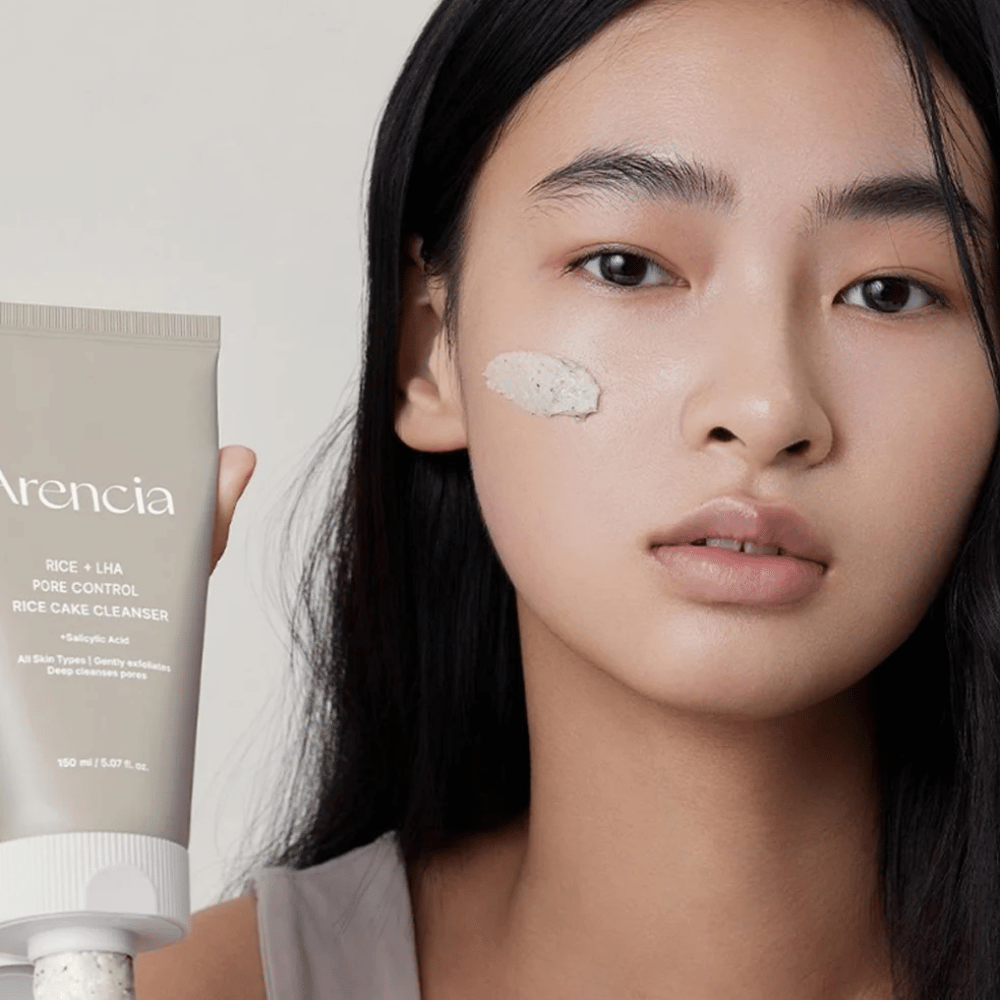 Modello che tiene Arencia Rice + LHA Pore Control Rice Cake Cleanser con prodotto applicato sulla guancia, illustra pulizia dei pori delicata ma efficace per l'uso quotidiano.
