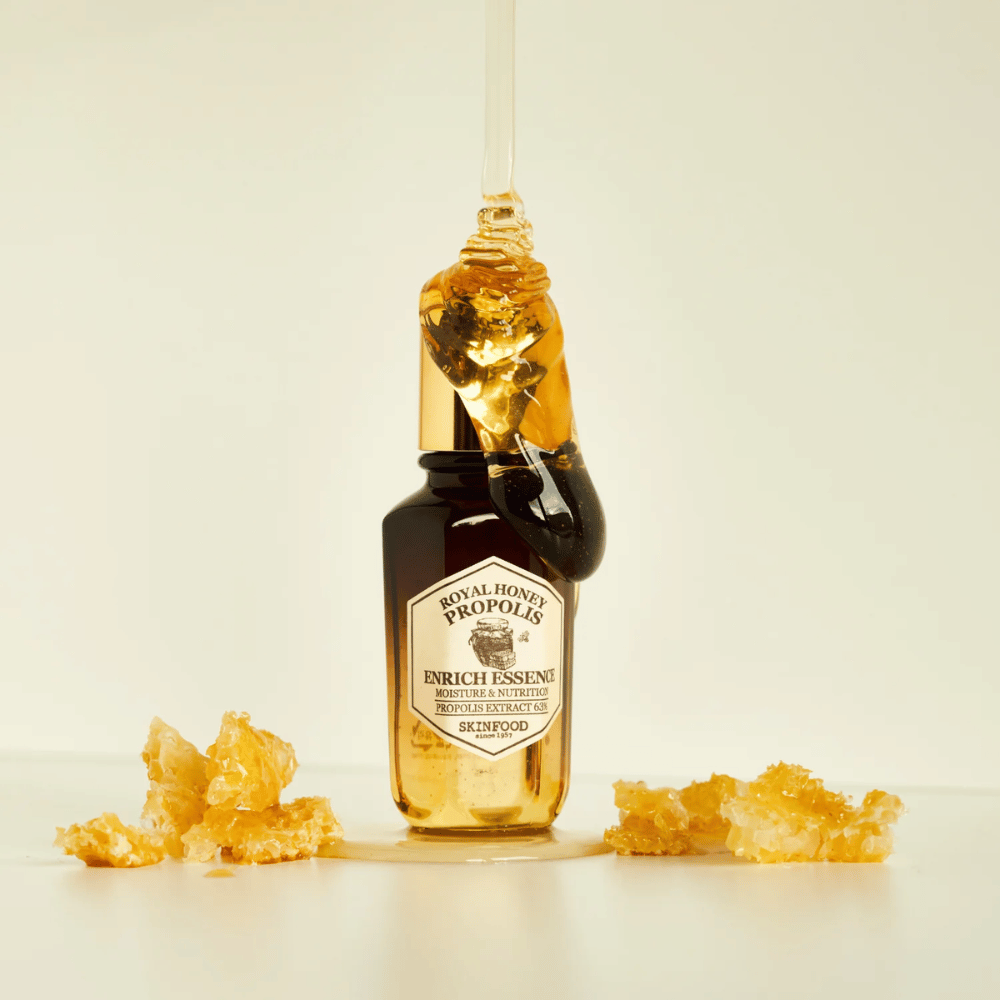 Immagine di una bottiglia di vetro con Royal Honey Propolis Enrich Essence da Skinfood. Una grande goccia di miele dorato scende dal tappo dorato della bottiglia. Intorno alla bottiglia ci sono piccoli pezzi di favo sparsi su una superficie chiara. L'essenza è commercializzata con un focus sull'umidità e la nutrizione, con il 63% di estratto di propoli.