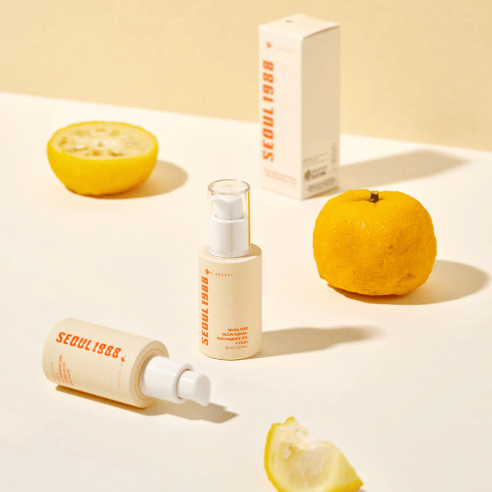 Diverse bottiglie e confezioni di Seoul 1988 Glow Serum disposte con yuja intere e tagliate a metà sullo sfondo.
