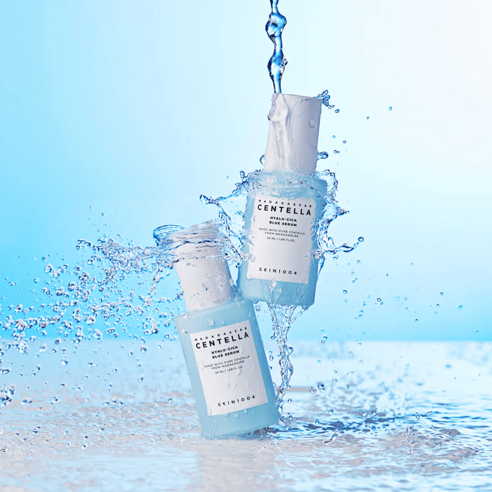 Due bottiglie di SKIN1004 Madagascar Centella Hyalu-Cica Blue Serum colpite da una cascata d'acqua su uno sfondo azzurro.