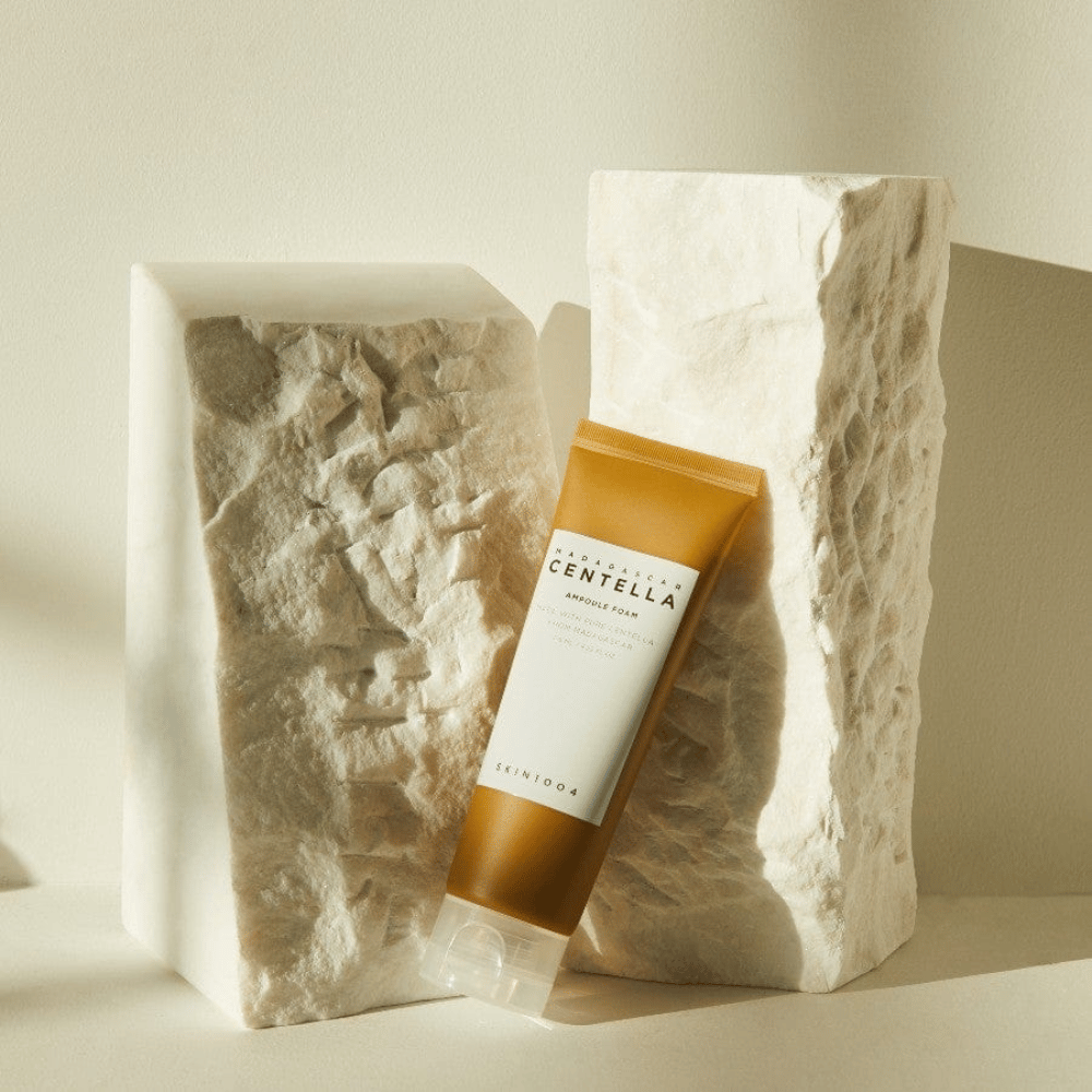SKIN1004 Madagascar Centella Ampoule Foam appoggiato contro blocchi di pietra chiara alla luce naturale del sole, con il suo design a tubo marrone-bianco e minimalismo elegante.