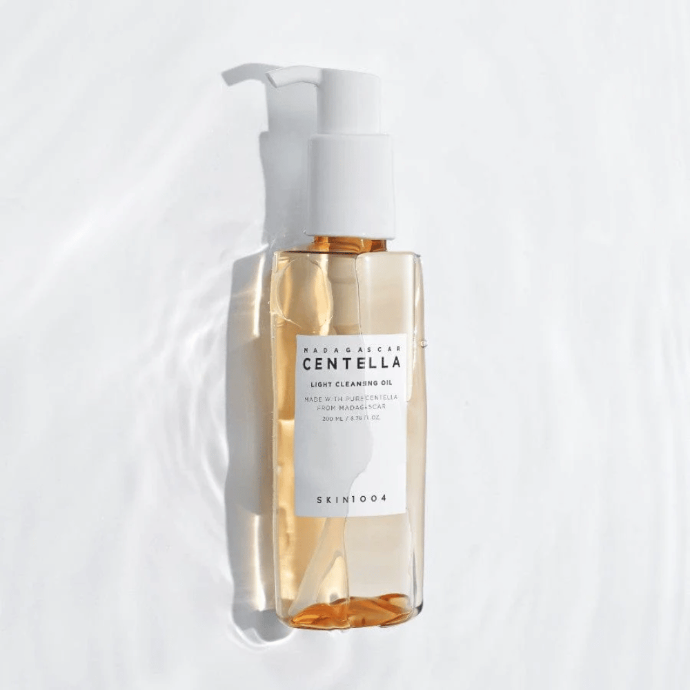SKIN1004 Madagascar Centella Light Cleansing Oil su sfondo bianco con morbidi riflessi d'acqua che mettono in risalto la bottiglia trasparente e il contenuto dorato.