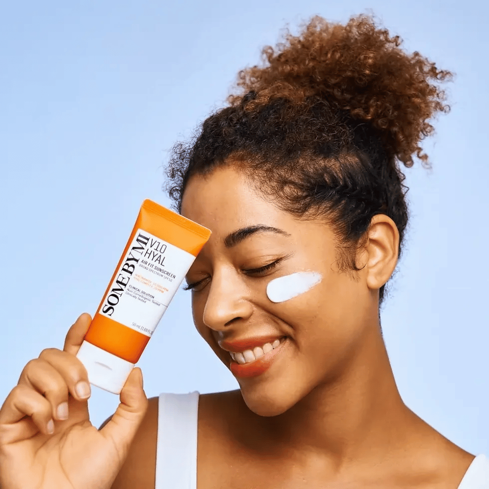 Una donna tiene in mano un tubetto di SOME BY MI V10 Hyal Air Fit Sunscreen e ha una macchia bianca di crema solare sulla guancia. Sorride felicemente e mostra chiaramente il prodotto, evidenziandone l'uso e i benefici per la cura della pelle e la protezione solare.