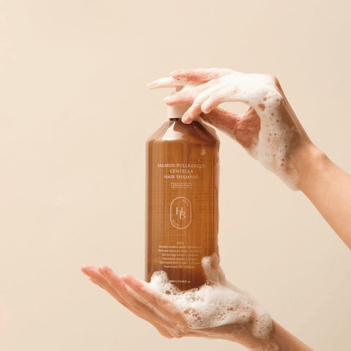 Heveblue Salmon Pullkeratin Centella Hair Shampoo in flacone marrone circondato da schiuma. Il prodotto è tenuto tra due mani e mette in risalto un'esperienza di cura dei capelli detergente e nutriente con abbondante schiuma.