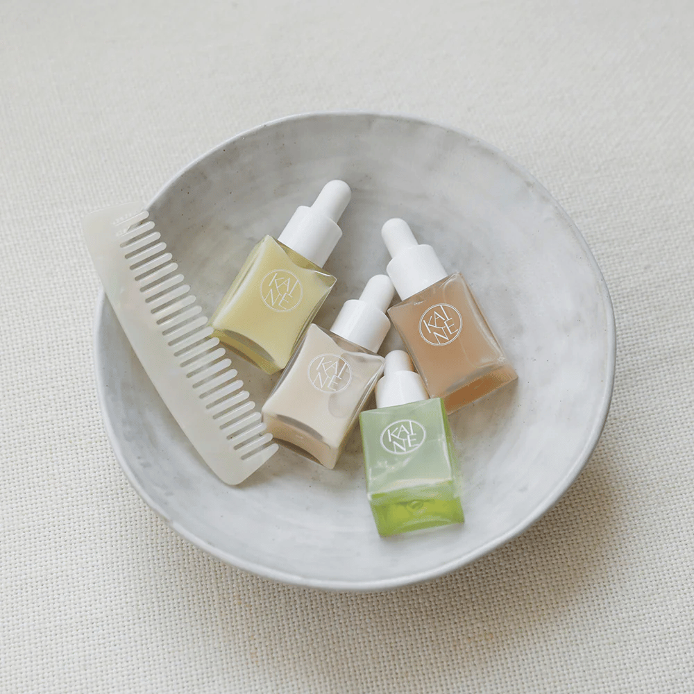 Quattro piccole bottiglie di siero di KAINÉ Serum Discovery Kit si trovano in una ciotola bianca accanto a un pettine, con uno sfondo naturale e calmo di tonalità beige.