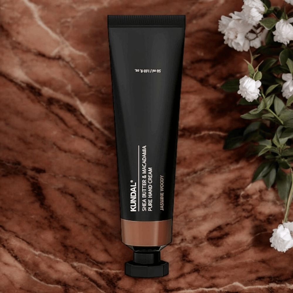 KUNDAL Shea Butter & Macadamia Pure Hand Cream Jasmine Woody posizionata su marmo con fiori – crema mani nutriente che cura le mani secche e offre un'elegante fragranza jasmine woody.