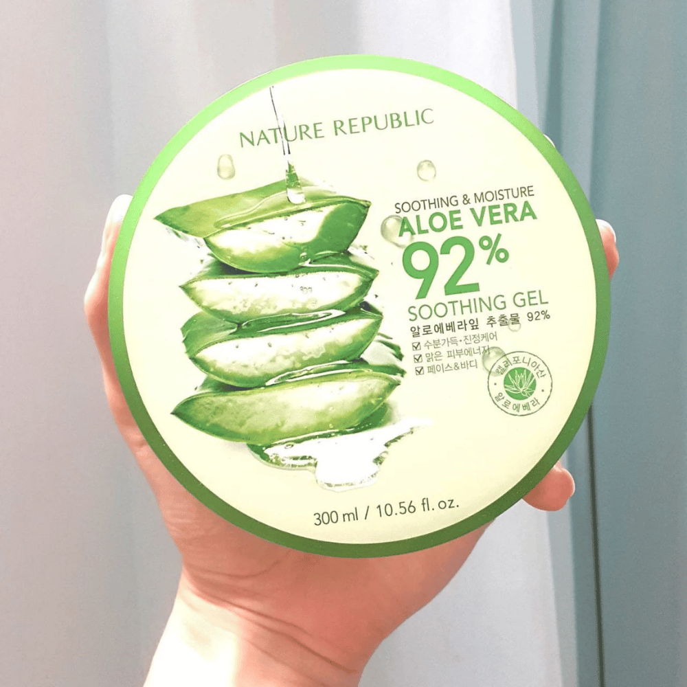 Una mano tiene un barattolo rotondo con Nature Republic Aloe Vera 92% Soothing Gel. Il barattolo è verde con un'immagine di aloe vera e testo che descrive le sue proprietà lenitive e idratanti. Il contenuto è di 300 ml.