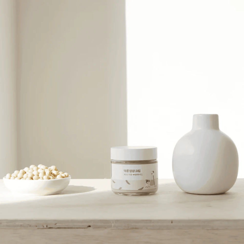 Mixsoon Soybean Nourishing Cream si trova su un tavolo di legno accanto a un vaso e fagioli di soia. Una crema idratante minimalista con ingredienti naturali.
