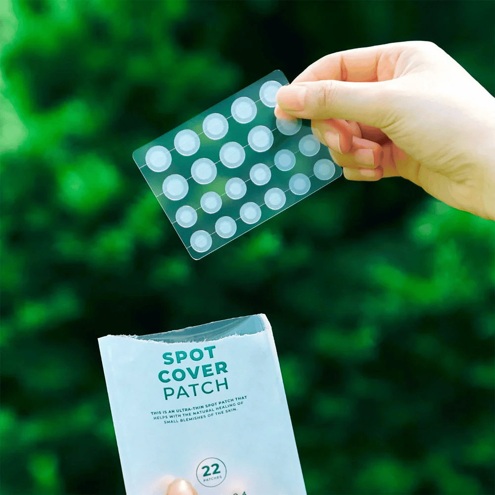 Una mano tiene un foglio con Spot Cover Patches, che sono trasparenti e arrotondati. Sullo sfondo si vede un ambiente verde sfocato. La confezione mostra una parte della confezione con il testo "SPOT COVER PATCH" e informazioni sul fatto che il prodotto contiene 22 cerotti che aiutano a guarire piccole aree della pelle.