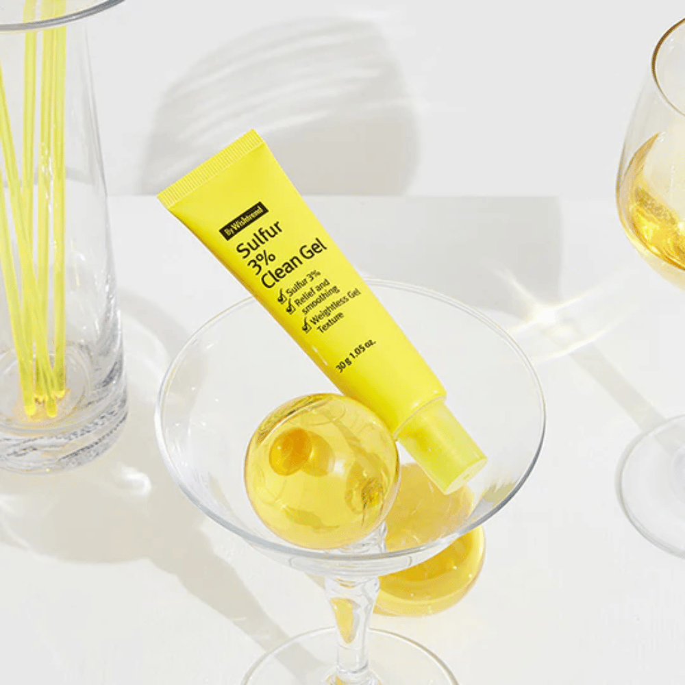 By Wishtrend Sulfur 3% Clean Gel in un tubo giallo posizionato in un bicchiere da cocktail con una sfera gialla, circondato da eleganti bicchieri su uno sfondo bianco.