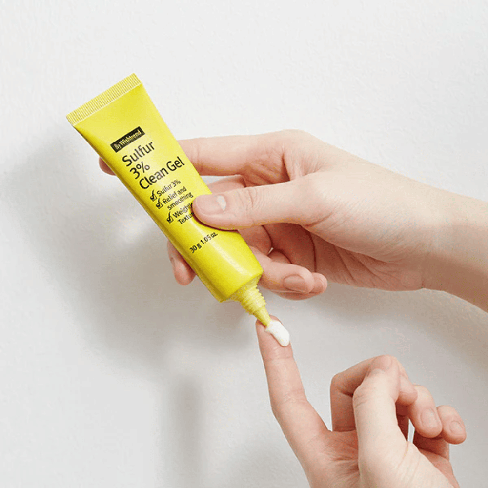 Primo piano di mani che applicano By Wishtrend Sulfur 3% Clean Gel, un tubo giallo con gel bianco, su uno sfondo bianco.