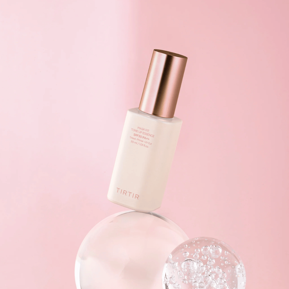 L'immagine mostra una bottiglia di "TIRTIR MASK FIT TONE UP ESSENCE" con SPF30 PA++ su uno sfondo rosa, che conferisce una sensazione di morbidezza e femminilità.