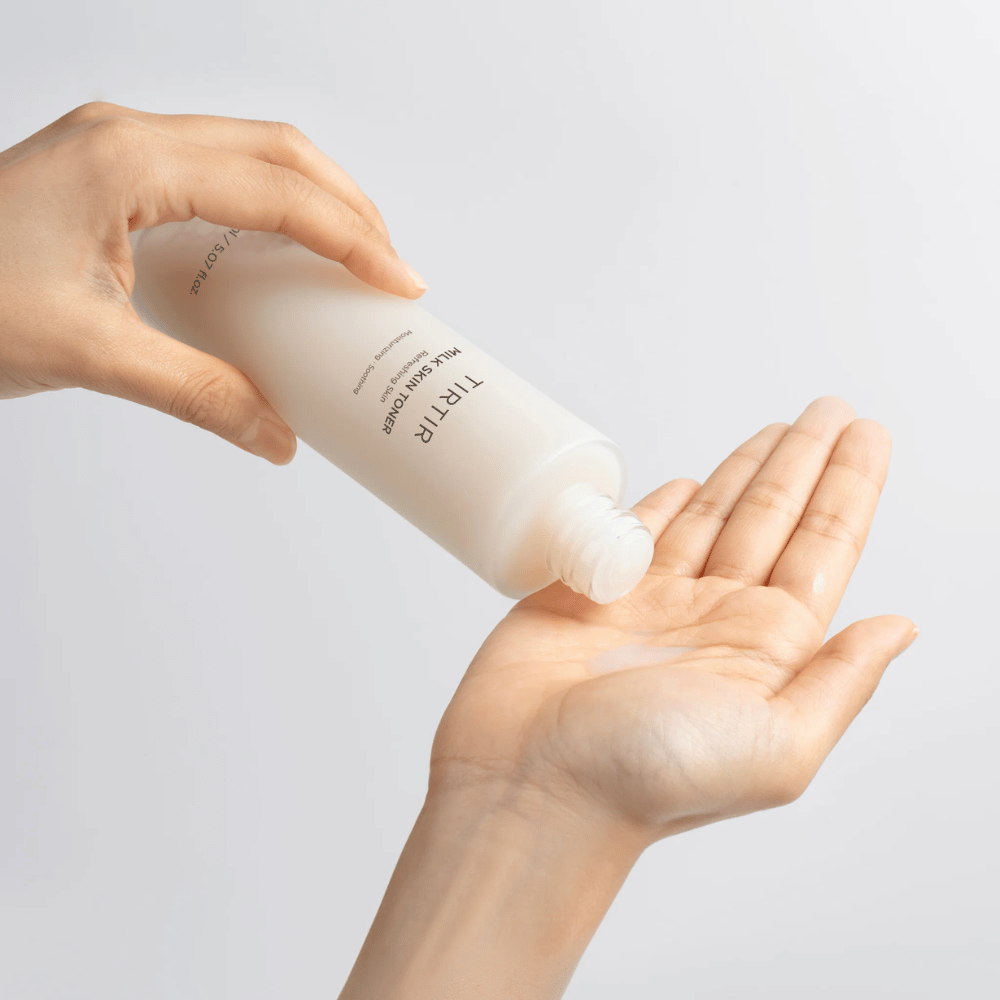 L'immagine mostra una persona che versa un liquido lattiginoso da una bottiglia etichettata "TIRTIR Milk Skin" sul palmo della mano. Il prodotto sembra essere un tonico per la pelle o una lozione idratante.