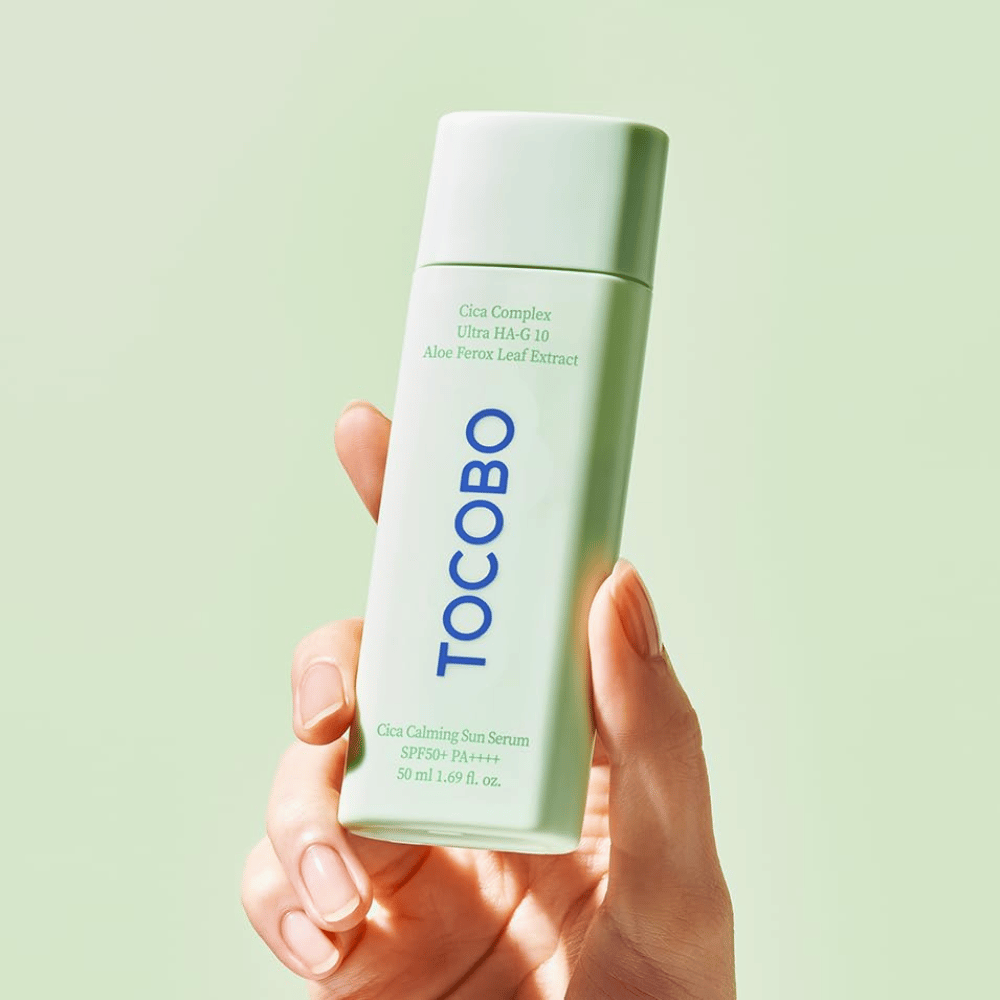 Mano che tiene TOCOBO Cica Calming Sun Serum con confezione verde menta e testo blu su sfondo verde chiaro.