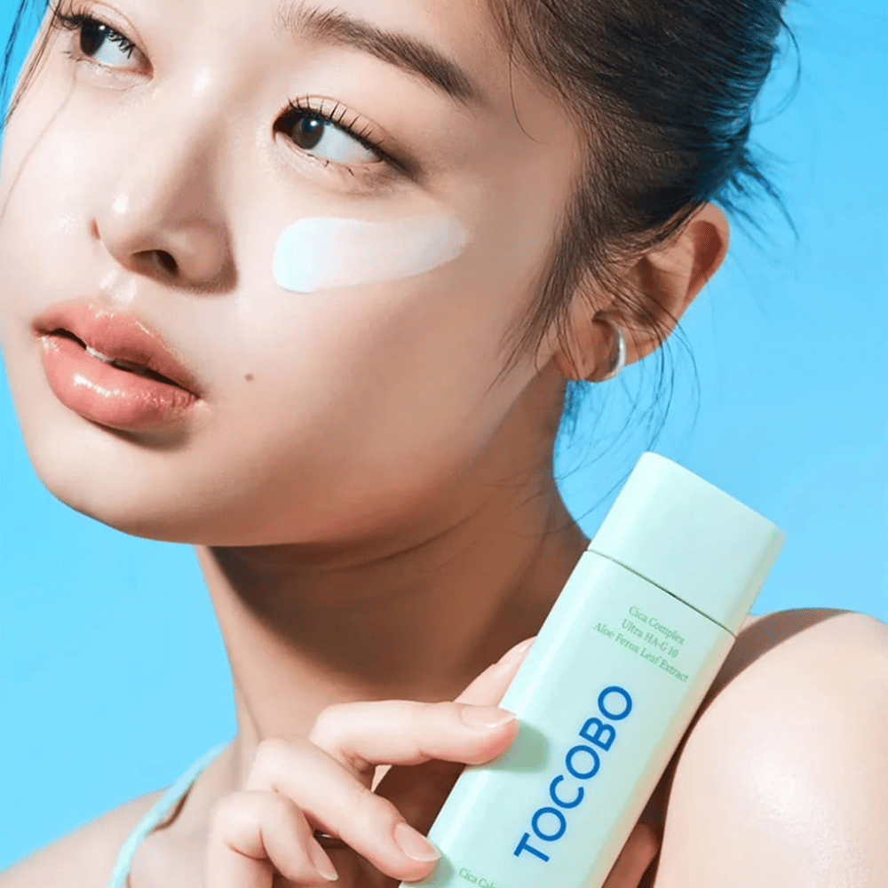Modello con crema solare sotto l'occhio tiene TOCOBO Cica Calming Sun Serum con confezione verde menta su sfondo blu.