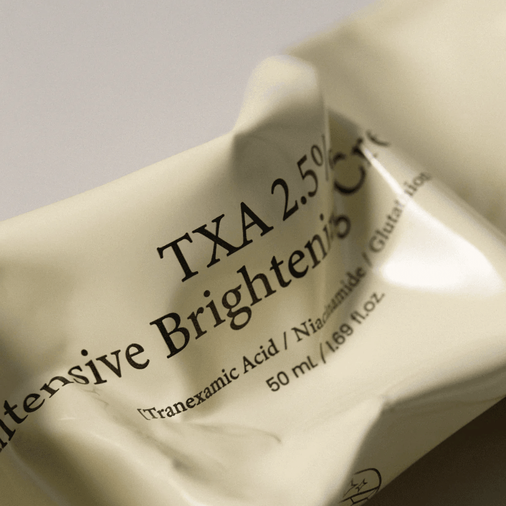 Primo piano di AXIS - Y TXA 2.5% Intensive Brightening Cream in cui il design morbido e minimalista del tubetto è al centro dell’attenzione. L’immagine mette in risalto il nome del prodotto, Tranexamic Acid e Niacinamide, nonché l’elegante confezione beige.