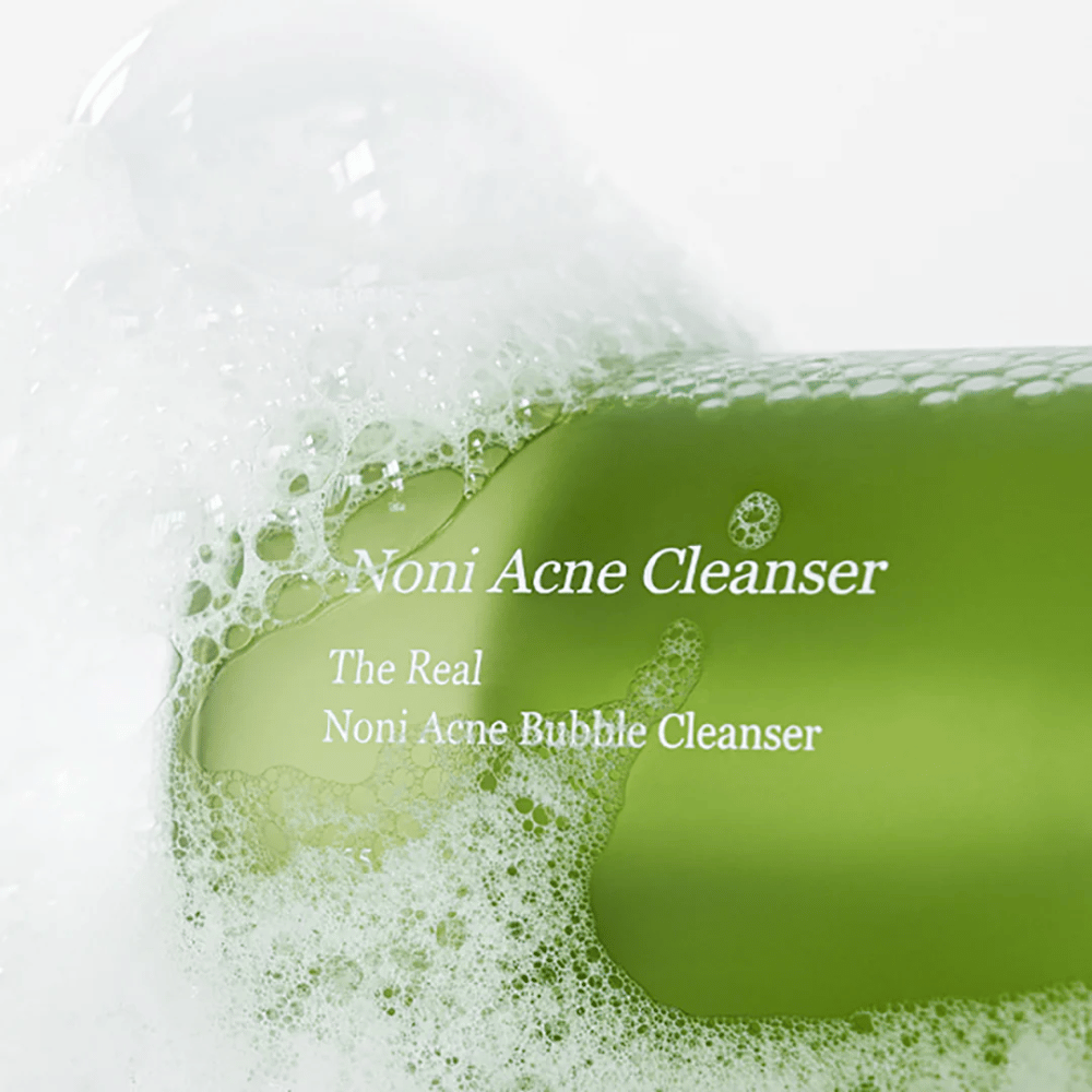 La bottiglia con The Real Noni Acne Bubble Cleanser è in primo piano e circondata da abbondante schiuma. Il colore verde fresco segnala ingredienti naturali, perfetti per pulire e combattere l'acne. Un prodotto ideale per una pelle pulita e chiara.