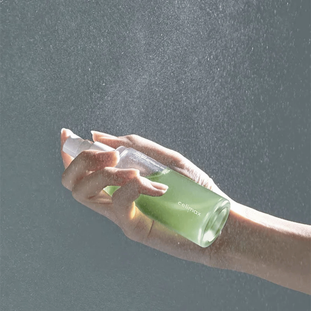 Una mano tiene Celimax Noni Mist spray e applica il prodotto con uno spruzzo fine e uniforme sulla pelle. La bottiglia è verde e il prodotto è commercializzato come "The Real Noni Energy Ampule Mist" per dare energia e idratazione alla pelle.