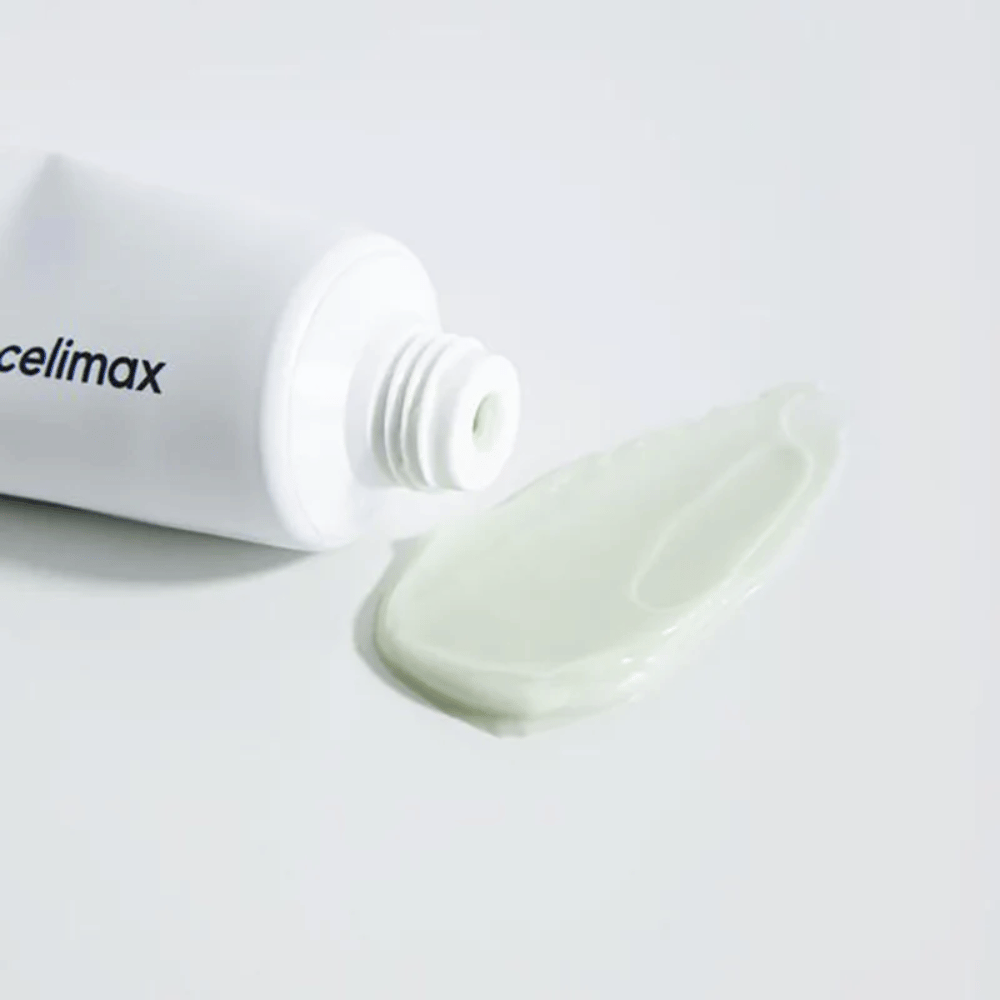 Celimax Noni Energy Repair Cream in un tubo bianco con una piccola quantità di crema verde chiaro spruzzata accanto al tubo su una superficie bianca. La crema è per nutrire e riparare la pelle.