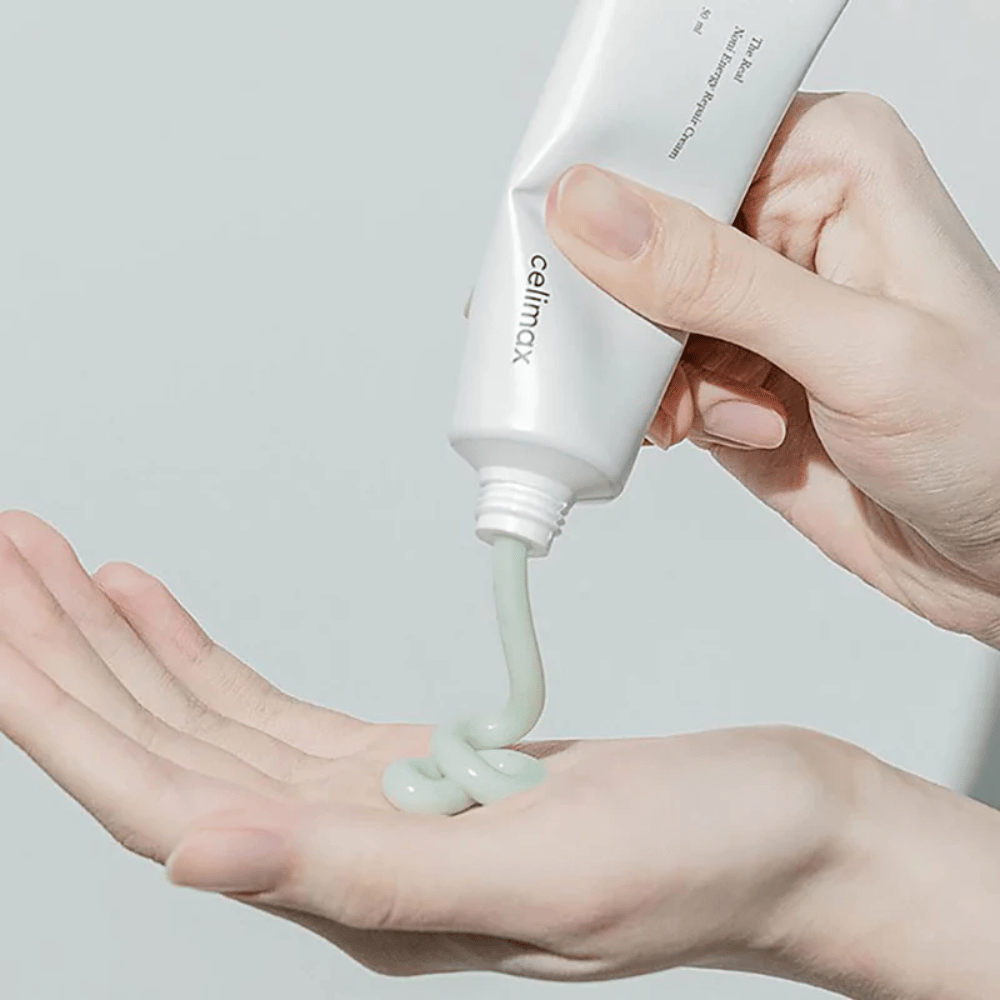 Una mano tiene un tubo bianco con Celimax Noni Energy Repair Cream, e la crema viene spremuta in un flusso uniforme sul palmo della mano. La crema è verde chiaro e viene commercializzata per nutrire e ripristinare la pelle.