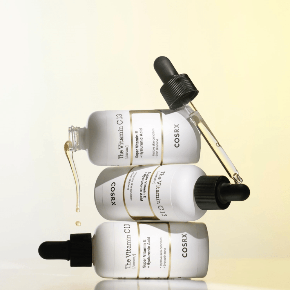 COSRX The Vitamin C 13 Serum con vitamina E e acido ialuronico, tre bottiglie con pipetta e gocce di siero alla vitamina C contro il tono della pelle irregolare e per luminosità.