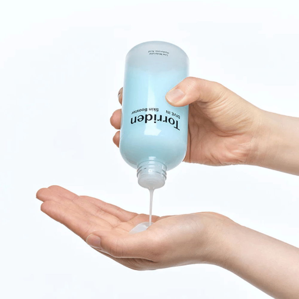 Mani che tengono una bottiglia di prodotto per la cura della pelle e versano un liquido trasparente nel palmo della mano. La bottiglia è etichettata con "Torriden - DIVE-IN Low Molecule Hyaluronic Acid Skin Booster".