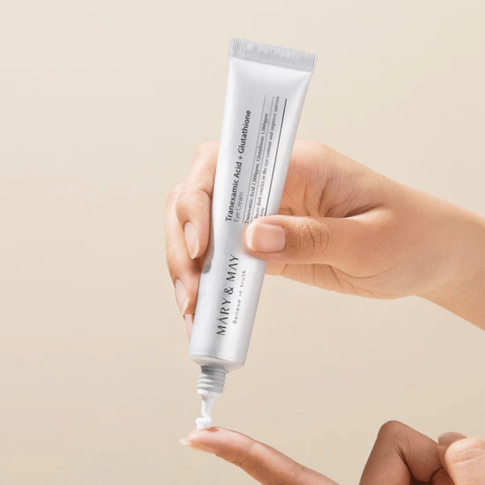 Una mano tiene un tubo di MARY & MAY "Tranexamic Acid + Glutathione Eye Cream". Il tubo è bianco e il design è pulito e semplice, il che evidenzia le proprietà calmanti e illuminanti del prodotto per l'area degli occhi. Il prodotto viene applicato delicatamente, con particolare attenzione alla precisione e all'efficacia.