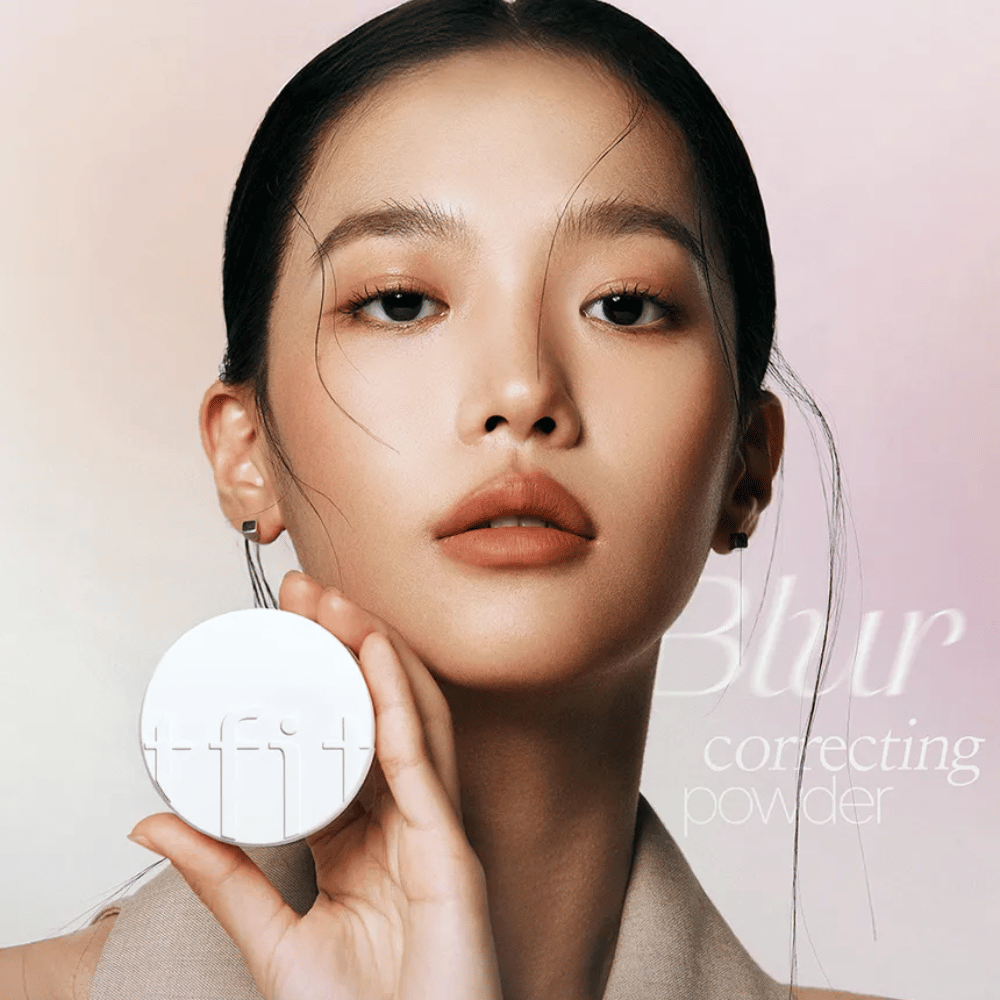 Donna con trucco impeccabile tiene una scatola rotonda bianca con il logo TFIT, con il testo "Blur Correcting Powder" sullo sfondo.