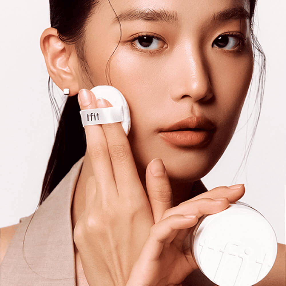 Donna applica TFIT Translucent Set Finishing Powder #02 Skin Beige con un puff, tenendo in mano una scatola di cipria bianca con il logo TFIT.