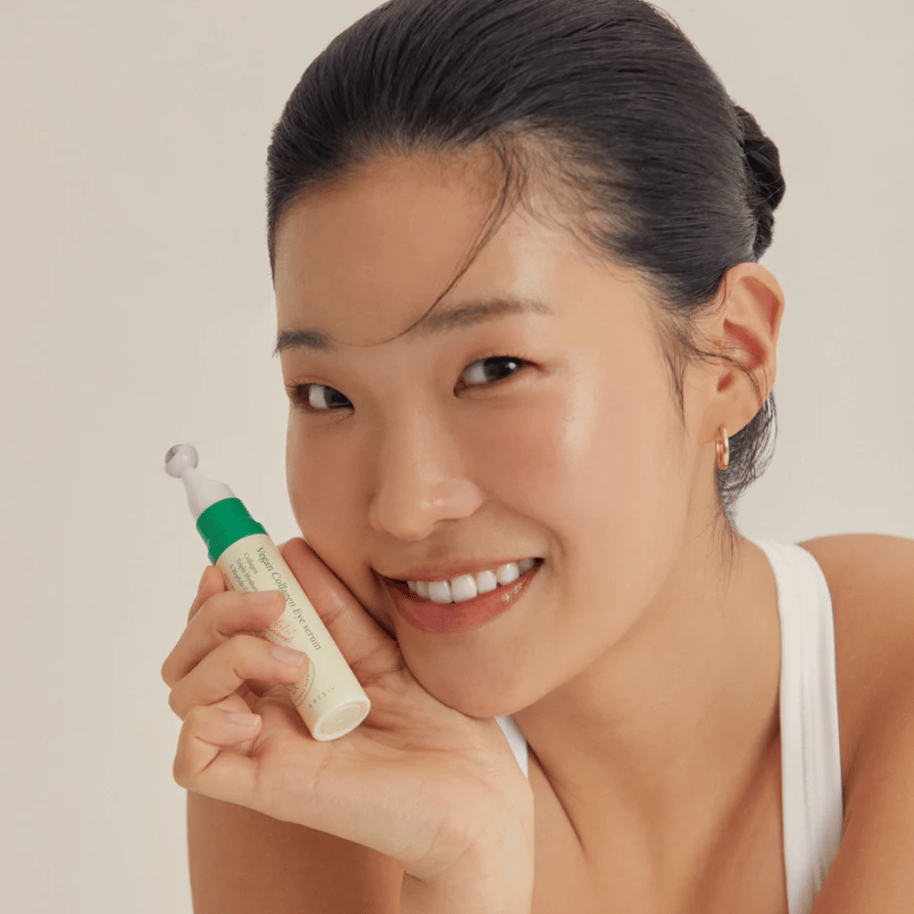 Una donna con i capelli raccolti e orecchini dorati tiene un AXIS-Y Vegan Collagen Eye Serum 10ml vicino al viso, mentre sorride dolcemente. Il siero è progettato per idratare e ridurre le rughe intorno agli occhi, appositamente formulato per pelli sensibili.