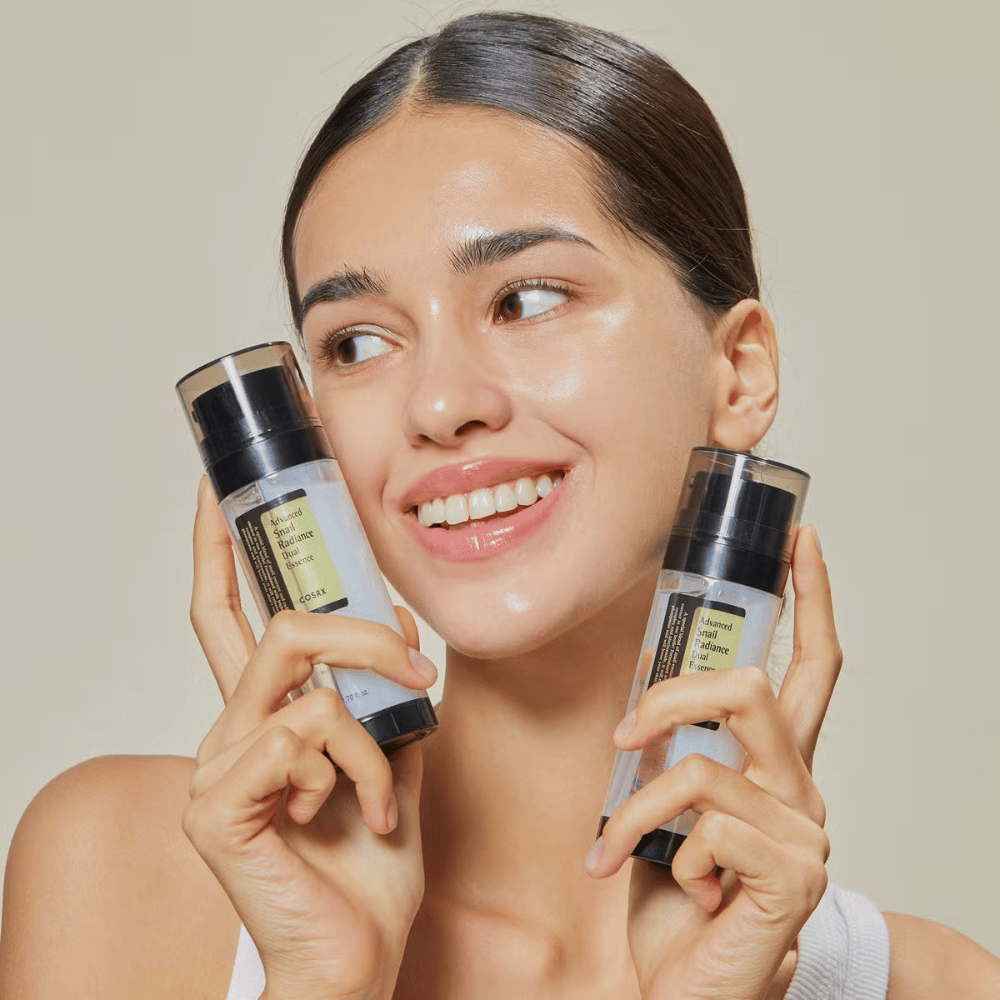Donna sorridente con pelle luminosa tiene due bottiglie COSRX Advanced Snail Radiance Dual Essence 80 ml vicino al viso su uno sfondo chiaro.