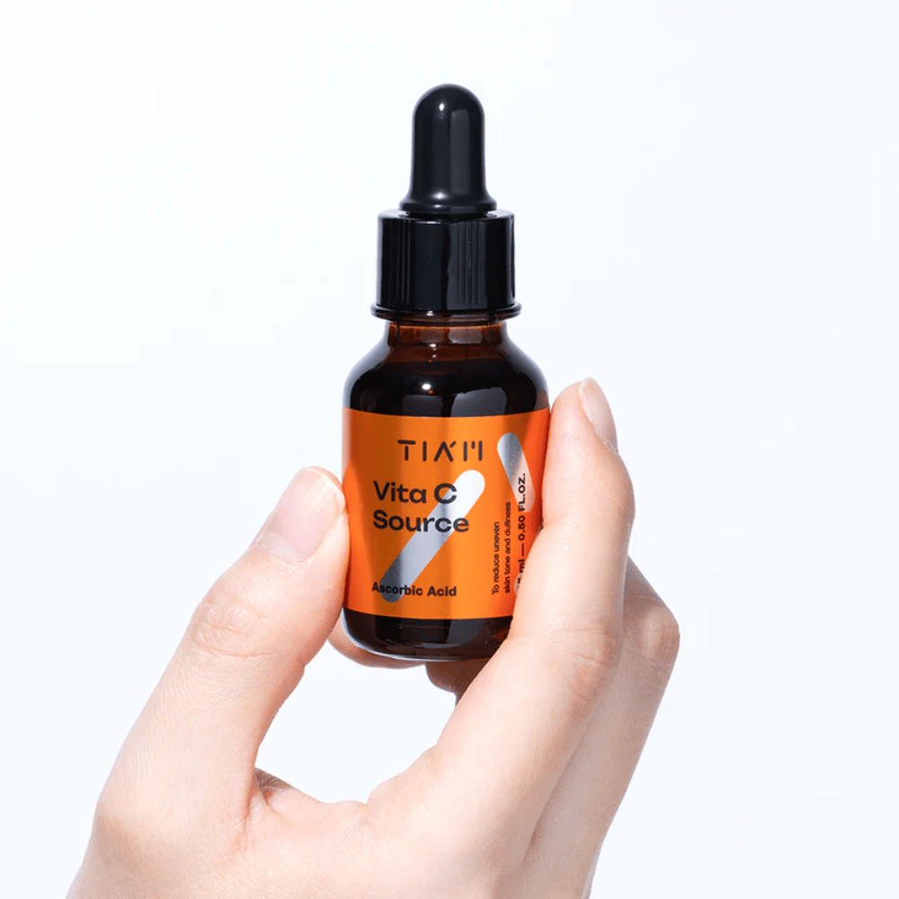 Una mano tiene TIA'M Vita C Source, un siero alla vitamina C in una bottiglia di vetro marrone con pipetta. Il prodotto è progettato per uniformare il tono della pelle e donare luminosità.