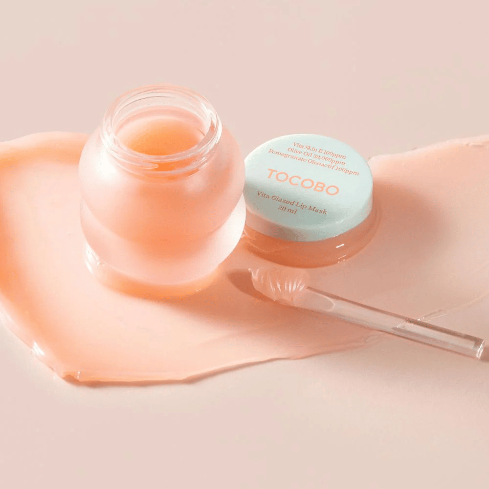 Un barattolo con "Vita Glazed Lip Mask" da Tocobo è mostrato. Il barattolo è trasparente con un coperchio turchese chiaro e contiene una maschera per labbra lucida e rosa. Una piccola spatola è accanto al barattolo, e una parte della maschera è sparsa sulla superficie. Il prodotto è progettato per idratare e curare le labbra.