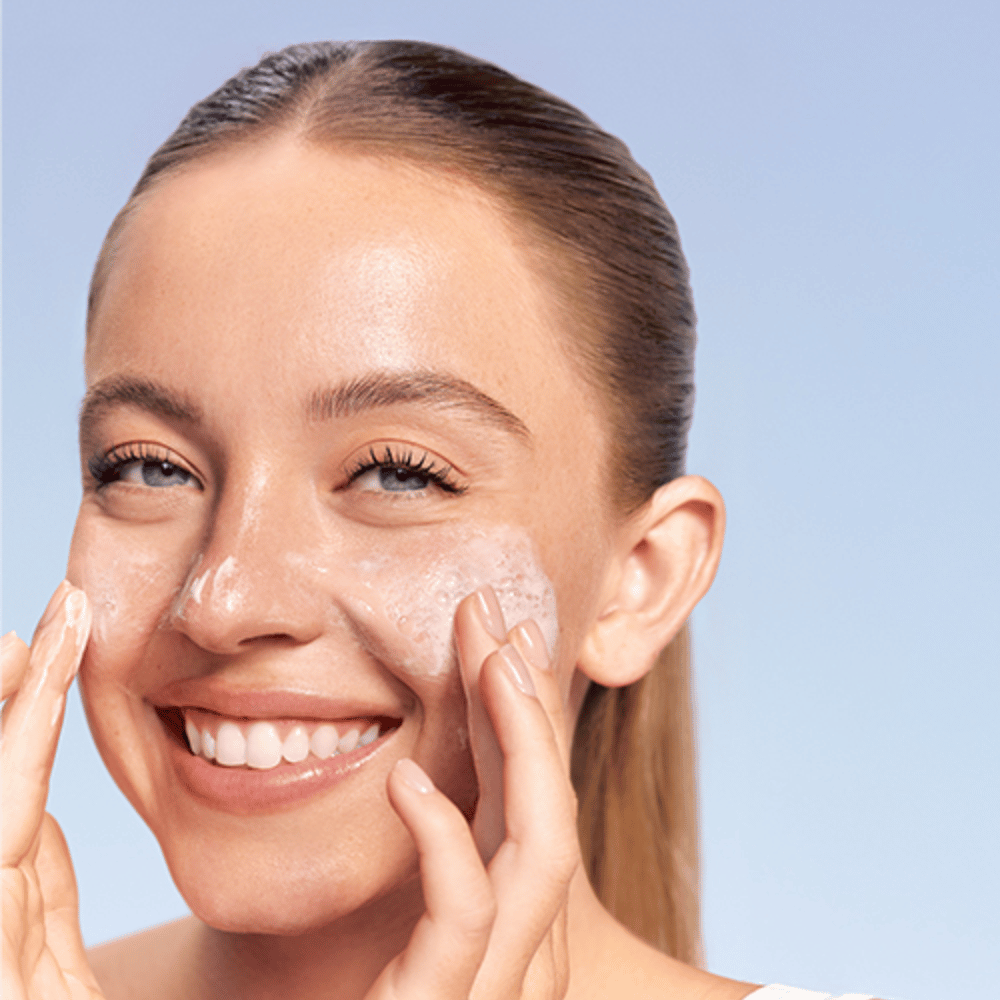 Donna sorridente applica LANEIGE Water Bank Gentle Gel Cleanser in forma schiumosa sulle guance, su sfondo blu.