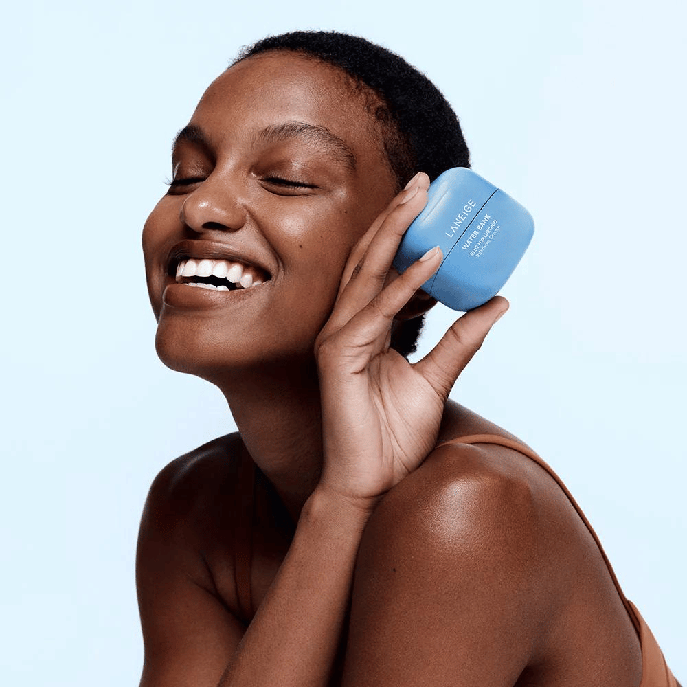 Donna sorridente tiene un barattolo blu di LANEIGE Water Bank Blue Hyaluronic Cream contro la guancia, con sfondo azzurro.