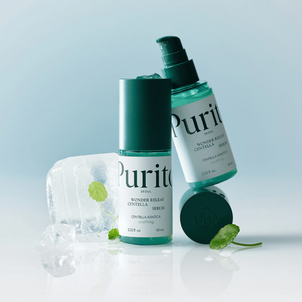 Due bottiglie di Purito Wonder Releaf Centella Serum, una in piedi e una sdraiata, circondate da cubetti di ghiaccio con foglie di centella all'interno e una foglia sciolta su una superficie azzurra.