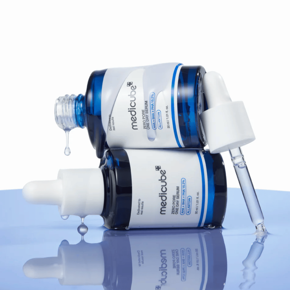 Due bottiglie blu di Medicube Zero Pore One Day Serum appoggiate l'una contro l'altra, con pipette e gocce di siero su una superficie blu riflettente.