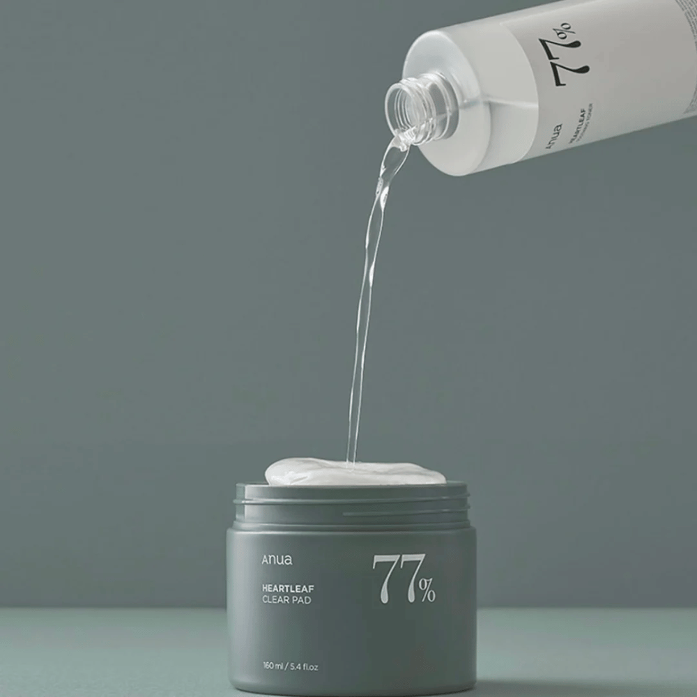 Una bottiglia trasparente versa un liquido in un barattolo grigio etichettato "anua HEARTLEAF 77% Clear Pad", che contiene una pila di dischetti per la cura della pelle bianchi. Lo sfondo è semplice e a tinta unita in una morbida tonalità di grigio.
